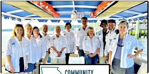 Group of people on a boat, wearing white shirts, smiling under a blue-striped canopy.